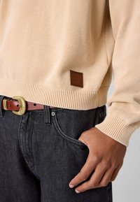 Beżowy dziergany sweter z prążkowanymi mankietami i dolnym brzegiem, z brązową skórzaną naszywką z logo. Noszony na ciemnoszarych dżinsach z brązowym paskiem.