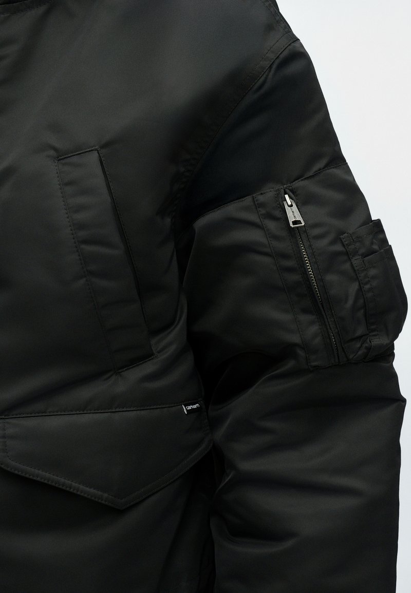 Schwarze gepolsterte Jacke mit einer glatten Gewebestruktur, Reißverschlusstaschen und einem sichtbaren Markenlabel am unteren Saum.