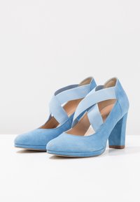 Chaussures à talons hauts en daim bleu avec un bout rond, dotées de brides élastiques croisées et d'un talon épais. Intérieur en cuir lisse.