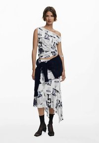 Vit asymmetrisk topp med grafiskt tidningsprint, kombinerad med en lång vit kjol med liknande mönster och en mörkblå stickad midjebindel.