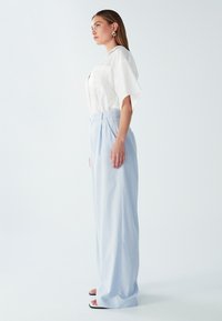 Bela satenska bluzica s kratkimi rokavi, v kombinaciji z svetlo modrimi, visokimi širokimi hlačami. Model nosi črne sandale in krožne uhane.