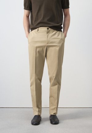 Bărbat purtând pantaloni bej croiți pe măsură, cu pliuri în față, cămașă maro închis cu mâneci scurte, pantofi negri slip-on, stând cu mâinile în buzunare.