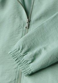 Veste zippée vert clair avec une texture lisse, des poignets élastiques et une fermeture éclair en métal, affichant un design simple et moderne.