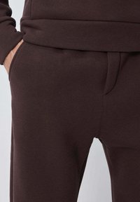 Pantalon de survêtement marron en tissu doux, avec un design fuselé, des poches et une ceinture élastique confortable. Texture lisse.