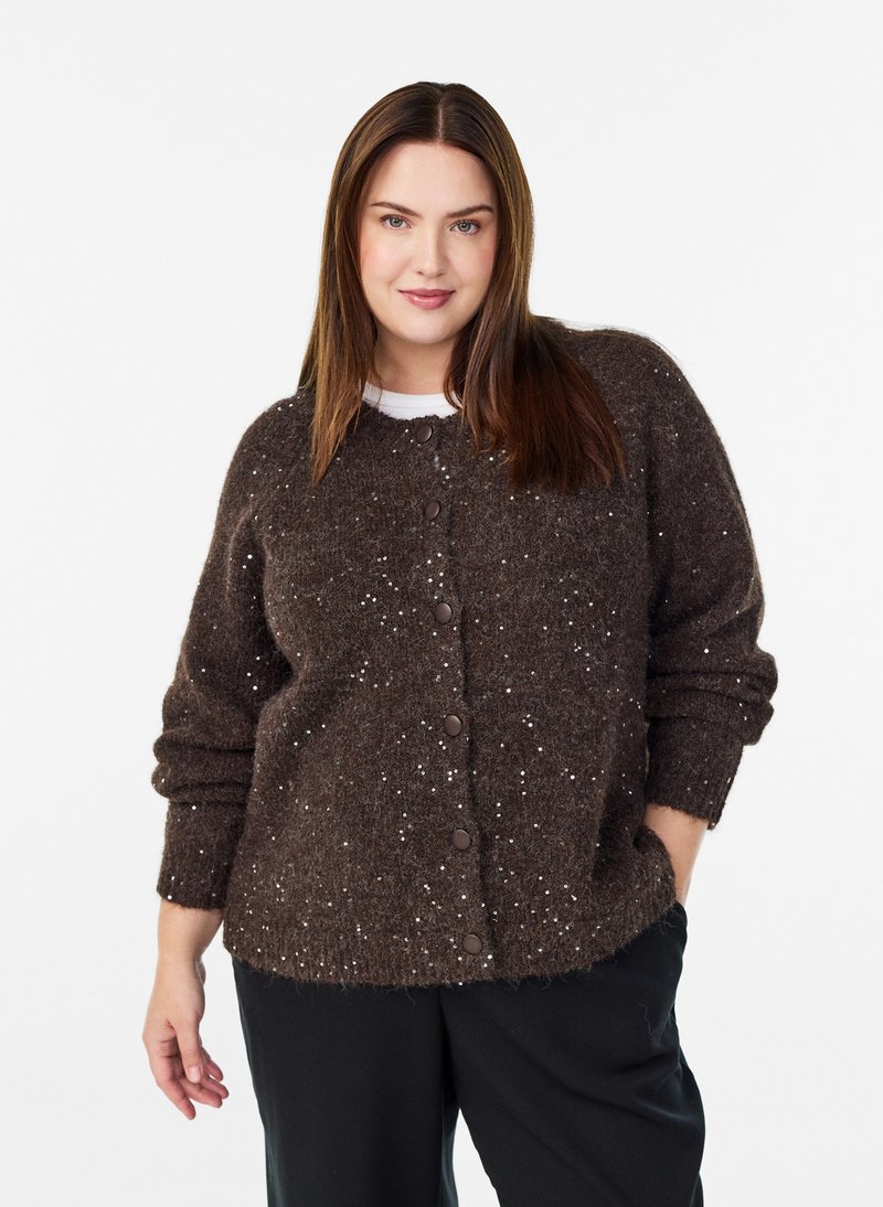 Donna con i capelli lunghi castani che indossa un maglione marrone scuro con bottoni e macchie bianche e pantaloni neri, in piedi su sfondo bianco.