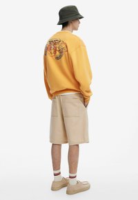 Gul sweatshirt med grafikdesign på ryggen, parret med beige shorts. Modellen bærer en grøn bøttehat og lyse sko med stribede sokker.