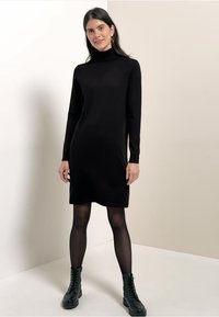 Svart långärmad polokragsklänning, figurnära, knä-lång, tillverkad av mjukt material. Kombinerad med svarta snörade ankelstövlar och genomskinliga strumpbyxor.