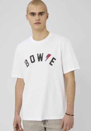 Jeune homme aux cheveux blonds courts portant un T-shirt blanc avec le texte "BOWIE" et un éclair rouge-bleu, pantalon gris clair, debout devant un fond uni.