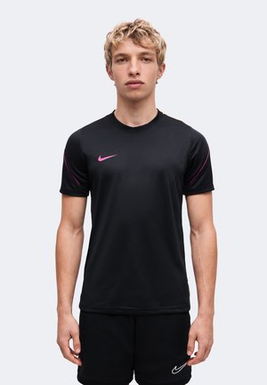 Jeune personne aux cheveux blonds courts et bouclés, portant une tenue de sport noire Nike avec un logo et des accents roses, debout devant un fond clair uni.