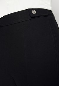 Černé kalhoty s hladkou tkaninou, které mají elegantní pas a kovový akcent na zapínání. Viditelné jemné švy.