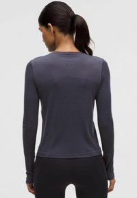 Tumedate soengiga naine, kellel on ponytail, seljas sobiv tumedashall spordisärk ja mustad retuusid, seisab seljaga lihtsa tausta poole.