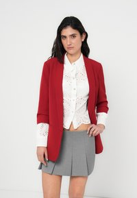 Κόκκινο σακάκι πάνω από μια λευκή μπλούζα με τρυπητό σχέδιο και κλείσιμο με κουμπιά, σε συνδυασμό με μια γκρι πλισέ φούστα. Υλικά με υφή και ξεχωριστές φόρμες και μοτίβα.