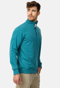 Sudadera de media cremallera en color verde azulado, hecha de una tela suave, con mangas largas, puños acanalados y una cremallera de color contrastante. Logo sutil en el pecho.