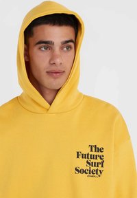 Žltá mikina s kapucňou z mäkkej textílie, s čiernym nápisom "The Future Surf Society" na ľavej strane hrudníka.