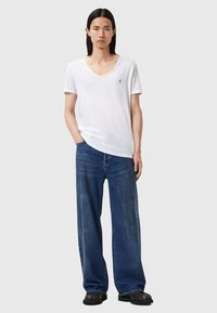 Witte t-shirt met ronde hals en korte mouwen, gecombineerd met een losse blauwe denim jeans. Het model staat met één hand in de zak, en draagt zwarte schoenen.