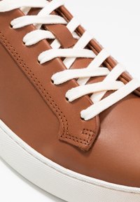 Bruna lädersneakers med vita snören, som har en rundad tå, sömnadsdetaljer och en texturerad gummisula.