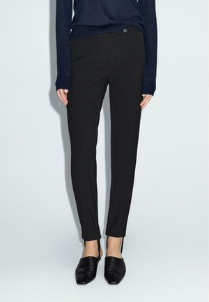 Pantaloni neri su misura a gamba slim, con un dettaglio di zip discreto all'orlo. Abbinati a un maglione in maglia blu scuro e scarpe nere slip-on.