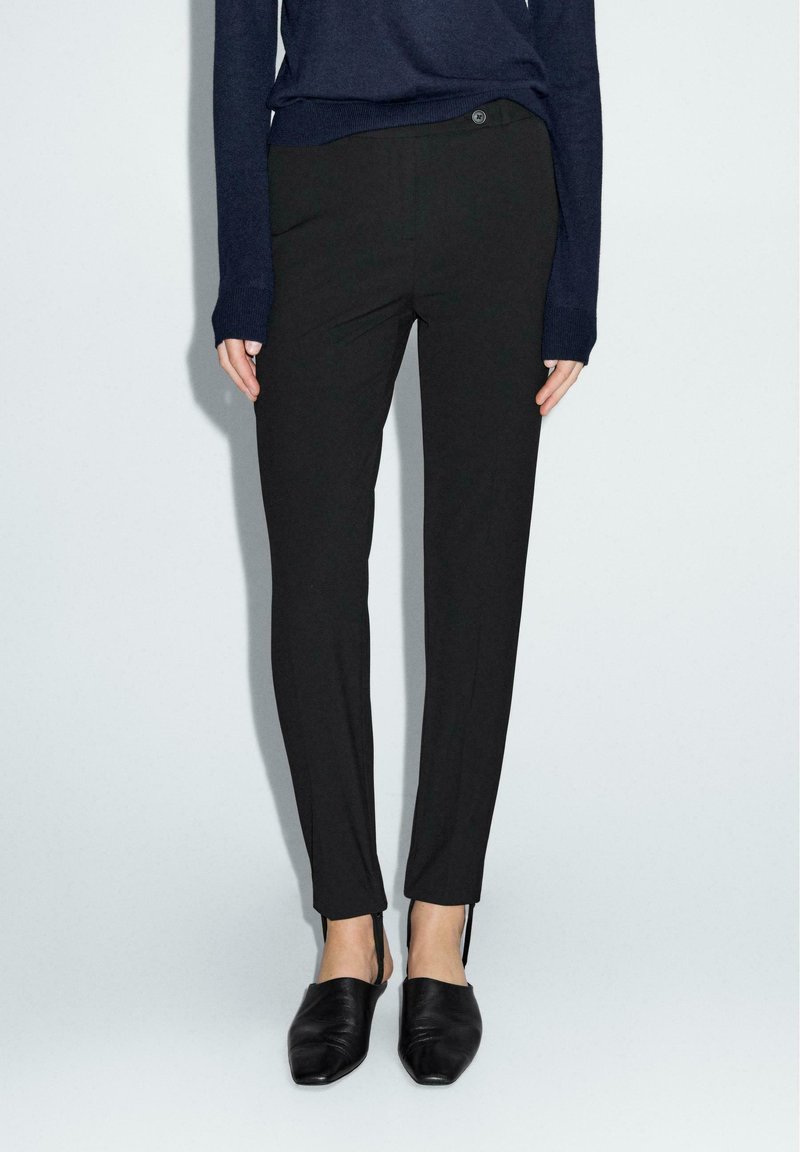 Pantaloni neri su misura a gamba slim, con un dettaglio di zip discreto all'orlo. Abbinati a un maglione in maglia blu scuro e scarpe nere slip-on.
