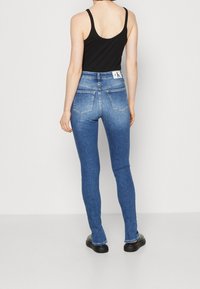 High-rise blue denim jeans with a slim fit, featuring a classic five-pocket design and stitching detail, worn with a black tank top.