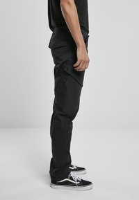 Schwarze Cargo-Hosen mit mehreren Seitentaschen, aus robustem Stoff, mit schmal zulaufendem Bein und einer glatten, matten Textur, kombiniert mit schwarzen Sneakers.
