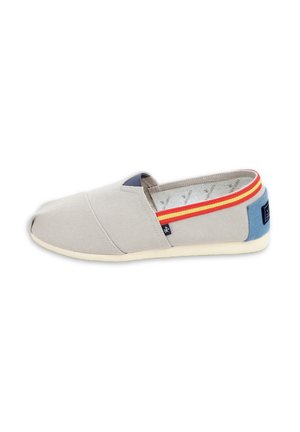 Zapato de lona gris claro sin cordones con ribete a rayas rojas y amarillas y parche azul en el talón sobre suela blanca, visto de lado.