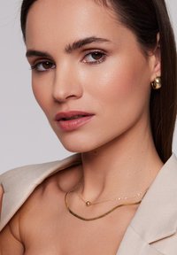 Złota biżuteria na modelce, przedstawiająca teksturowany, falisty naszyjnik z okrągłym akcentem oraz dużymi złotymi kolczykami w kształcie koła, umieszczona na neutralnym tle.