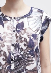 Blusa a maniche corte in tessuto leggero con stampa floreale in toni viola e grigi. Include una chiusura con bottoni davanti e una tasca sul petto.
