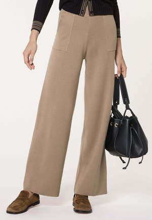 Femme portant un pantalon beige à jambes larges, des mocassins en daim marron, un haut noir, tenant un sac à main en cuir noir avec fermeture à cordon.