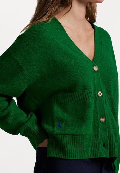 Grønn strikket cardigan med V-hals, ribbestrikkede mansjetter og to frontlommer. Har gullknapper og en blå brodert logo på lomma.
