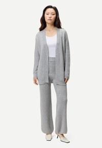 Frau steht, trägt eine hellgraue, strukturierte Strickjacke und passende, weit geschnittene Hosen, ein weißes Oberteil und weiße Schuhe mit Absatz.