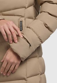 Beige pufferjack met een gewatteerd ontwerp. Close-up van een hand die een ritszak op de mouw aanpast. Zachte, gladde stof textuur.