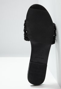 Semelle plate noire de sandale avec une bande de roulement texturée, des brides supérieures tressées et une indication de la taille imprimée sur la semelle intérieure sur fond blanc.