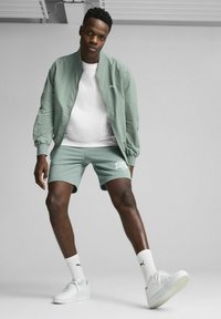 Mintgrøn bomberjakke over en hvid t-shirt og matchende shorts. Har et lille logo på jakken, hvide sokker og hvide sneakers.