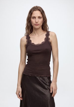 Vrouw met golvend haar die een donkerbruine geribde tanktop met kanten rand draagt en een zwarte leren rok, staand tegen een effen lichte achtergrond.