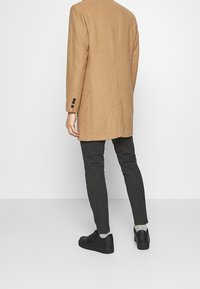 Lungo cappotto di lana beige con taglio dritto, tre bottoni sui polsini, abbinato a pantaloni scuri a righe sottili e sneakers nere, visto da dietro.