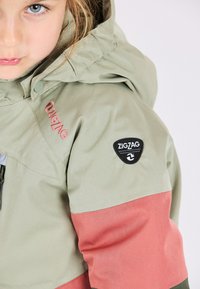 Chaqueta infantil verde y rosa con capucha, que presenta un exterior texturizado, un parche con el logo en zigzag y un panel de color contrastante a lo largo de las mangas.