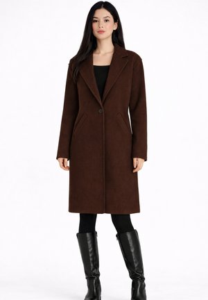 Frau trägt einen knielangen braunen Wollmantel, ein schwarzes Oberteil, schwarze Leggings und schwarze kniehohe Lederstiefel, steht vor weißem Hintergrund.