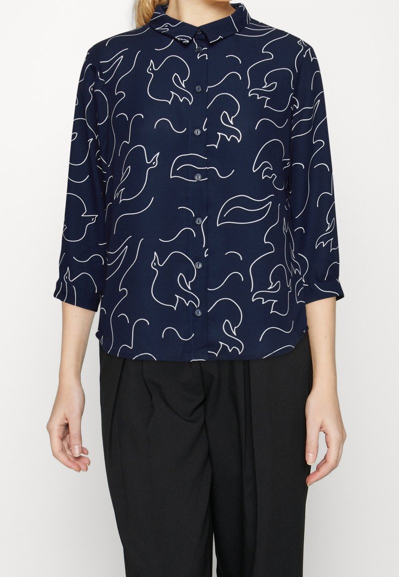 Femme portant une chemise boutonnée bleu marine avec des motifs abstraits d'oiseaux blancs et de vagues, associée à un pantalon noir ample.