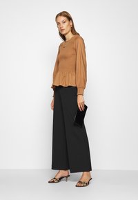 Rjava smockana peplum bluza z dolgimi rokavi, uparjena z širokimi črnimi hlačami. Model drži črno torbico in nosi leopardaste petke.