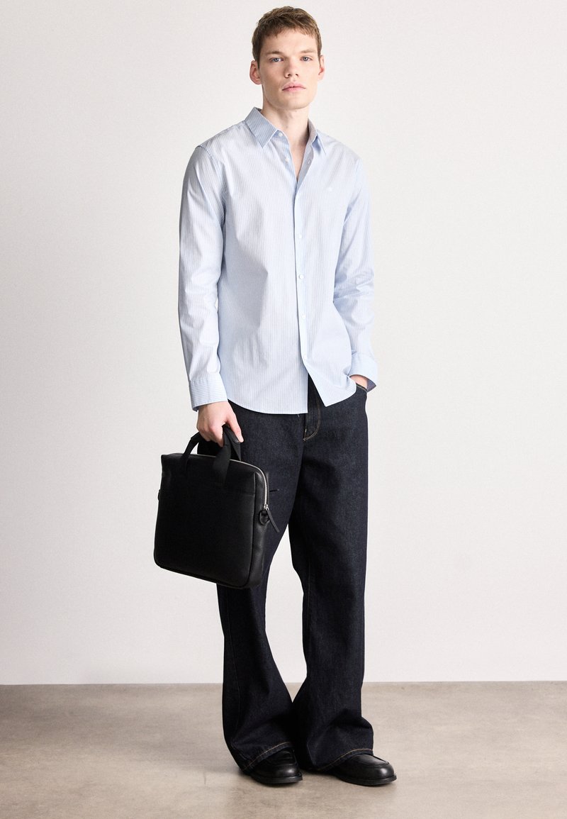 Hellblau gestreiftes Hemd mit Knöpfen, dunkle Jeans mit weitem Bein und schwarze Lederschuhe. Hält eine schwarze Ledertasche mit zwei Griffen.