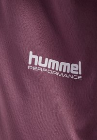 Nahaufnahme eines violetten, strukturierten Stoffes mit weißem "hummel PERFORMANCE"-Logo, das auf der Oberfläche aufgedruckt ist.
