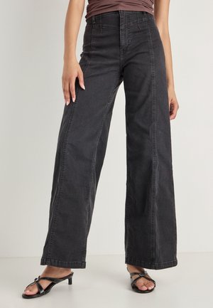 Pantalons en denim noirs à jambes larges avec une taille haute, présentant des détails de couture à l'avant et une texture lisse. Assortis à des sandales noires à lanières.