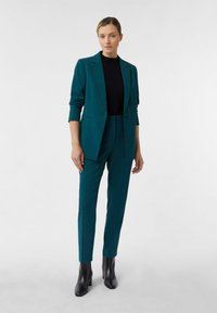 Frau in einem türkisfarbenen Anzug mit Blazer, passenden Hosen, schwarzem Oberteil und schwarzen Knöchelstiefeln vor einem schlichten weißen Hintergrund.