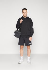 Black hoodie with drawstring, gray shorts, and white sneakers. Holds a black bag with a textured surface and white logo. Simple design.