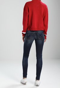Suéter rojo de gran tamaño y jeans oscuros ajustados con detalles desgastados en los bolsillos traseros, combinados con zapatillas blancas.