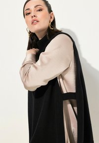 Maglione nero senza maniche in maglia sopra una camicia lunga rosa satinata a maniche lunghe, con collo alto e ritagli laterali per un contrasto di texture.