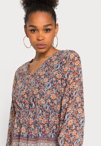 Vestido floral com fundo azul-marinho e flores laranja e brancas. Mangas longas, decote em V e detalhe de cintura franzida com barra estampada.