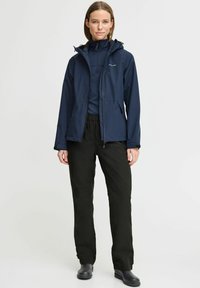 Marineblau wasserdichte Jacke mit Reißverschluss, verstellbarer Kapuze und zwei Seitentaschen, kombiniert mit schwarzen wasserdichten Hosen und robustem schwarzen Schuhwerk.