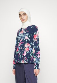 Top floral de mangas longas em azul marinho com flores rosas e verdes. Tecido suave com um corte solto e textura franzida nos punhos. Combinado com calças escuras.