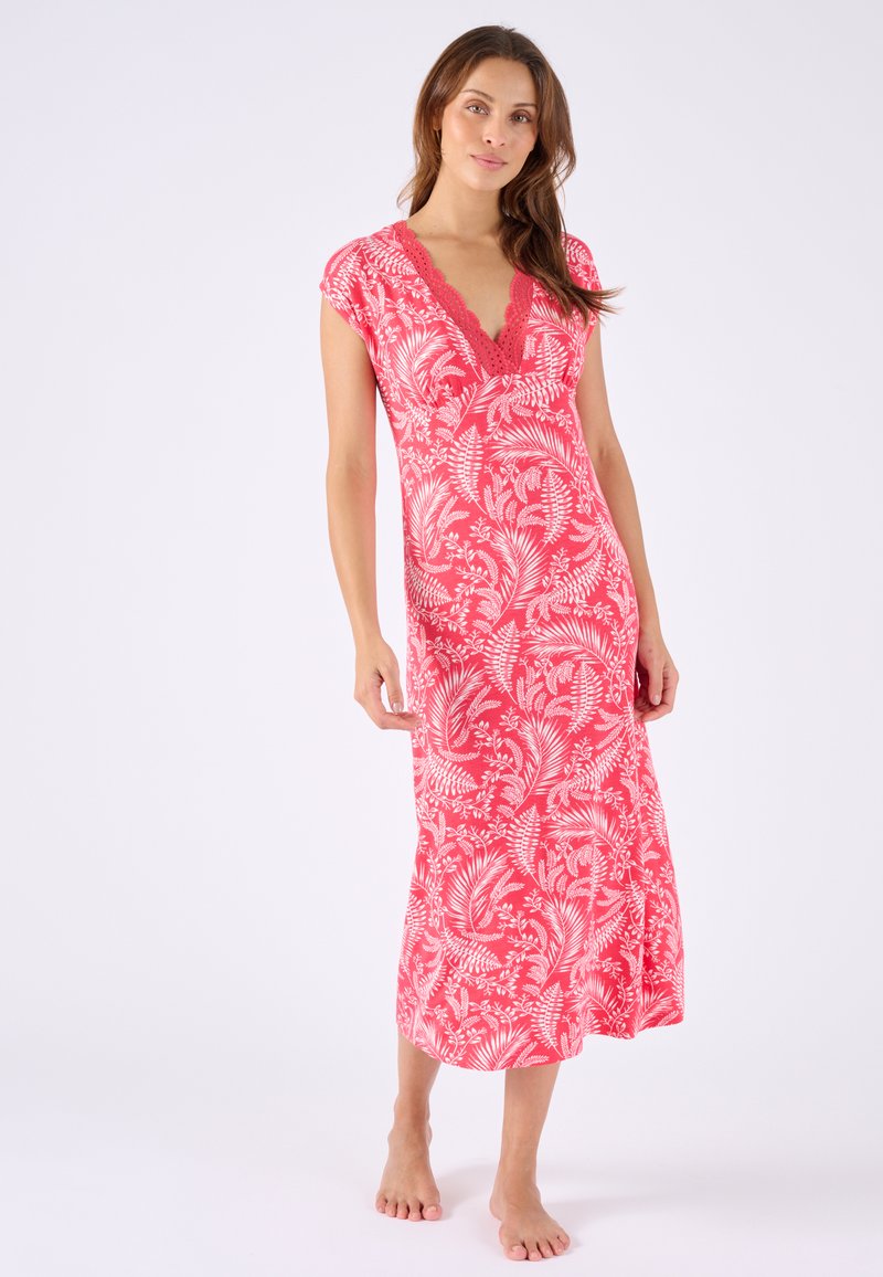 Femme debout pieds nus portant une robe midi corail avec des motifs de feuilles tropicales blanches et un décolleté festonné.